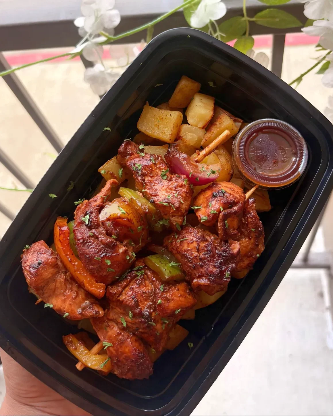 A container of chicken kebabs with Cajun roasted potatoes and a cup of BBQ sauce, held up on a balcony